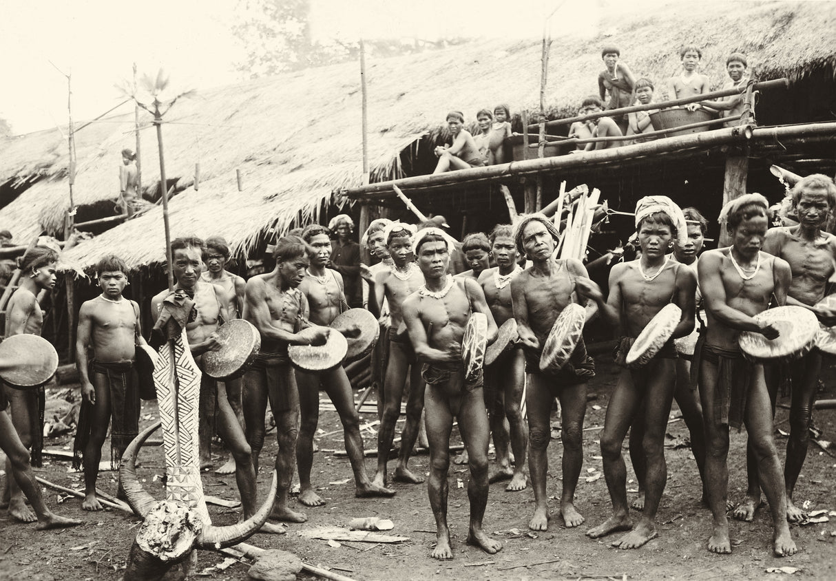 Archival photo of Dayak men drumming during a ceremony in front of a longhouse, central Borneo
