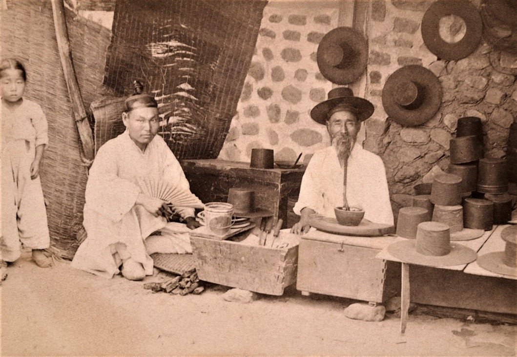 Sepia photograph from 1901 of two Korean artisans crafting traditional gat hats, seated in a workshop surrounded by hat molds, tools, and finished hats on a stone wall.