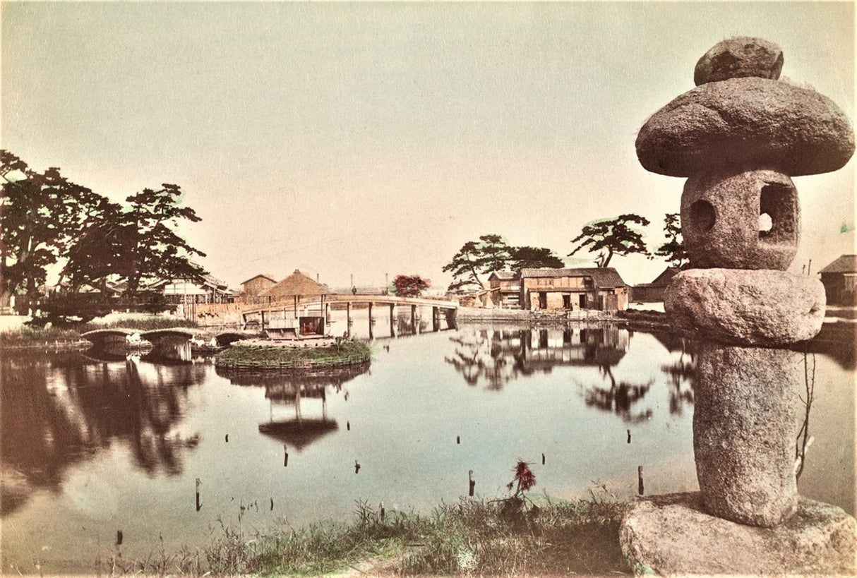 Hand-colored Meiji-era photograph of a traditional Japanese garden featuring a stone lantern, wooden bridge, pond, and surrounding architecture.