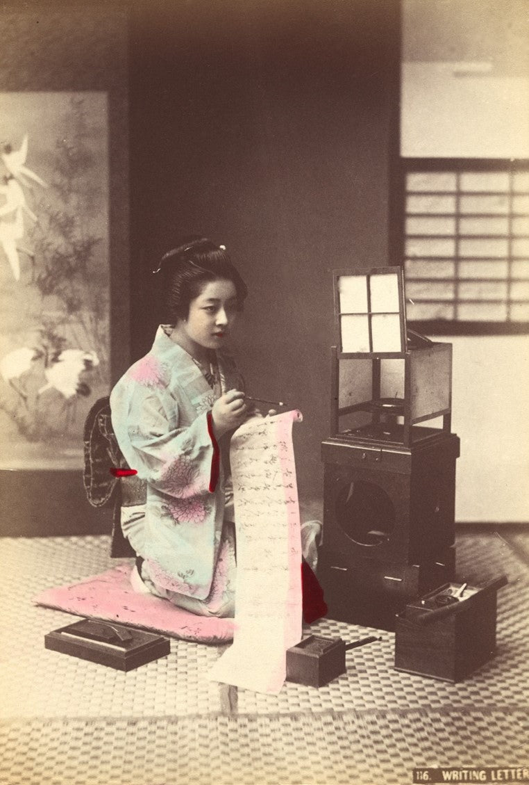Hand-colored Meiji-era photograph of a Japanese woman in a blue kimono writing a letter, seated in a traditional interior with lantern, screens, and calligraphy tools.