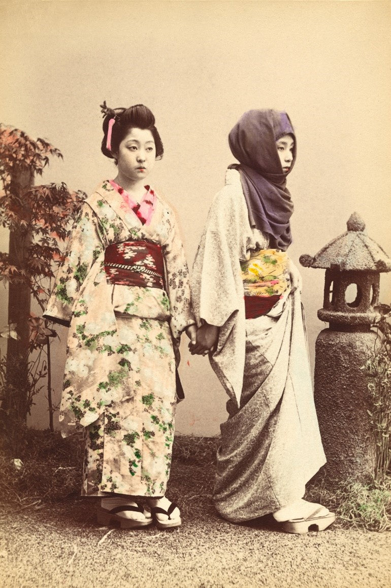 Hand-colored Meiji-era photograph of two Japanese women holding hands, dressed in kimono, walking beside a stone lantern.