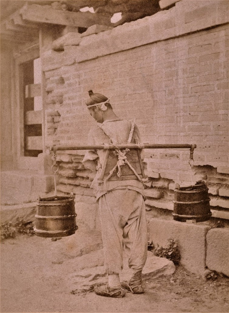Sepia photograph from 1901 showing a Korean man carrying water with a shoulder yoke and wooden buckets, dressed in traditional hanbok and walking past a stone wall.