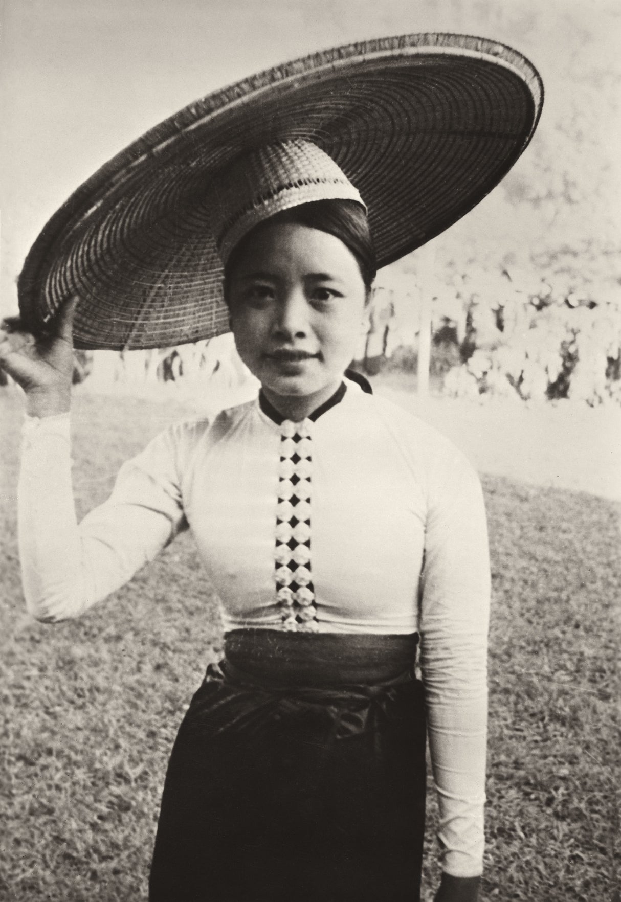 Black-and-white photo of a Vietnamese woman in traditional dress holding a large bamboo hat, 1950s