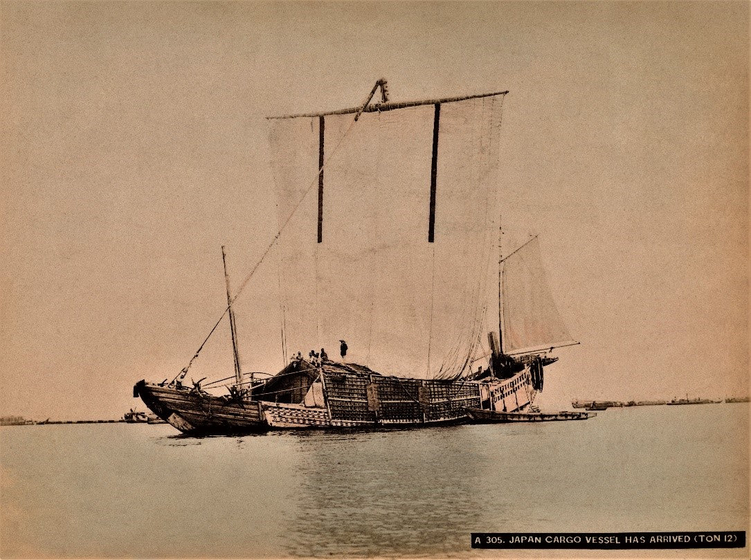 Hand-colored photograph of a traditional Japanese cargo ship with large sails, sailing on calm waters during the Meiji era.