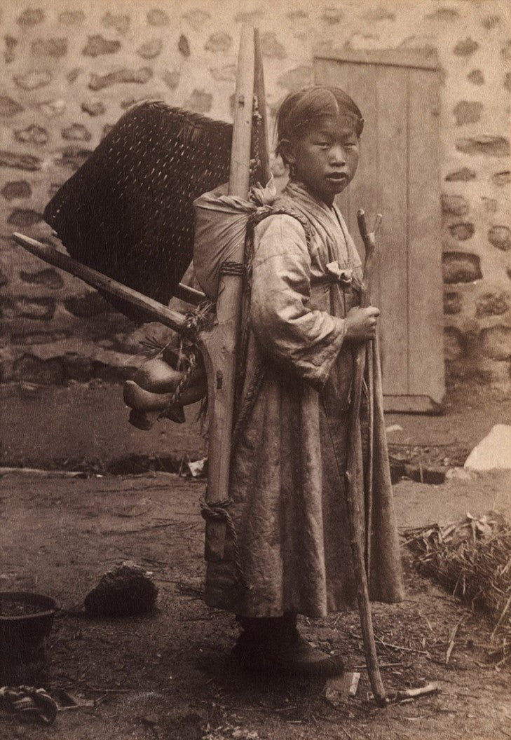 Sepia portrait of a young Korean figure wearing a traditional overcoat and carrying a bundle, photographed in a rustic courtyard during the early Korean Empire.