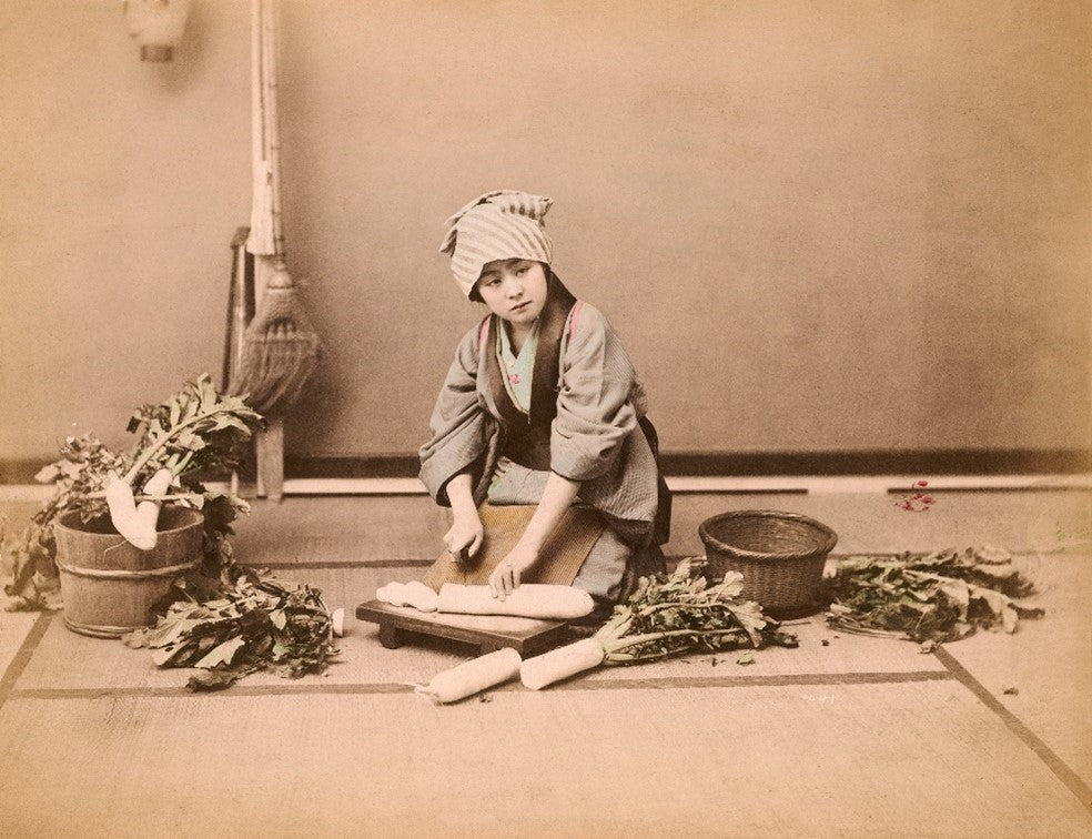 Hand-colored Meiji-era photograph of a Japanese woman slicing daikon radish in a traditional setting