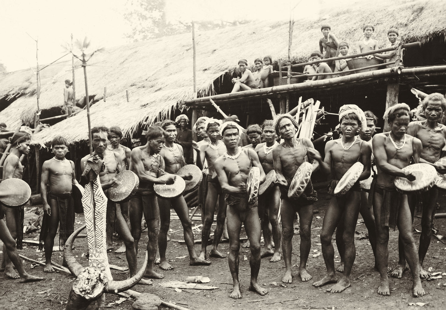 Archival photo of Dayak men drumming during a ceremony in front of a longhouse, central Borneo