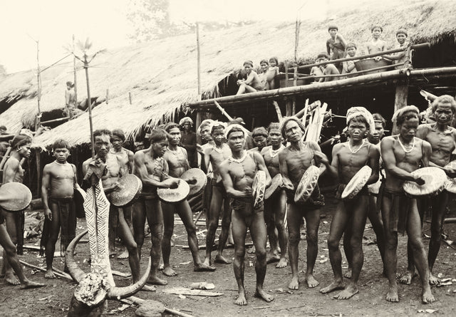 Archival photo of Dayak men drumming during a ceremony in front of a longhouse, central Borneo