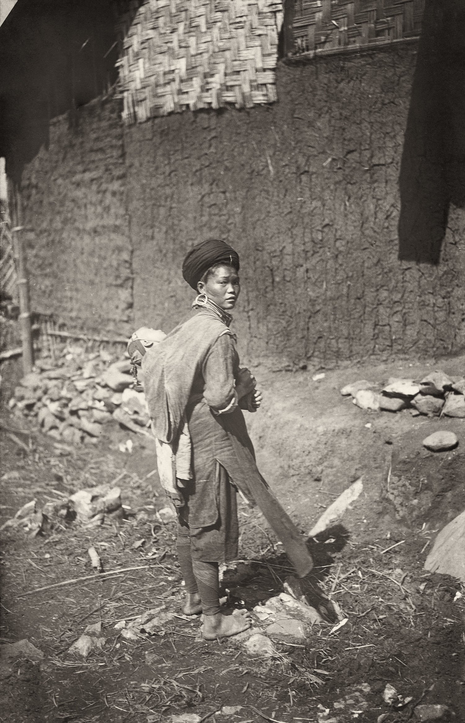 Woman in traditional dress with baby in cloth carrier, standing outside a thatched mud house