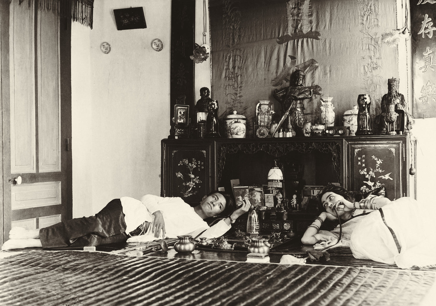 Sepia photograph of a man and woman reclining in an opium den, with ornate decor and Chinese-style furnishings