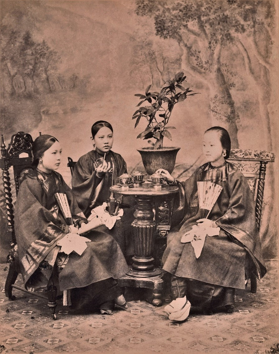 Sepia-toned 1901 studio portrait of three Chinese women in traditional silk garments seated around a tea table, engaging in a formal tea-drinking ritual in Hong Kong.
