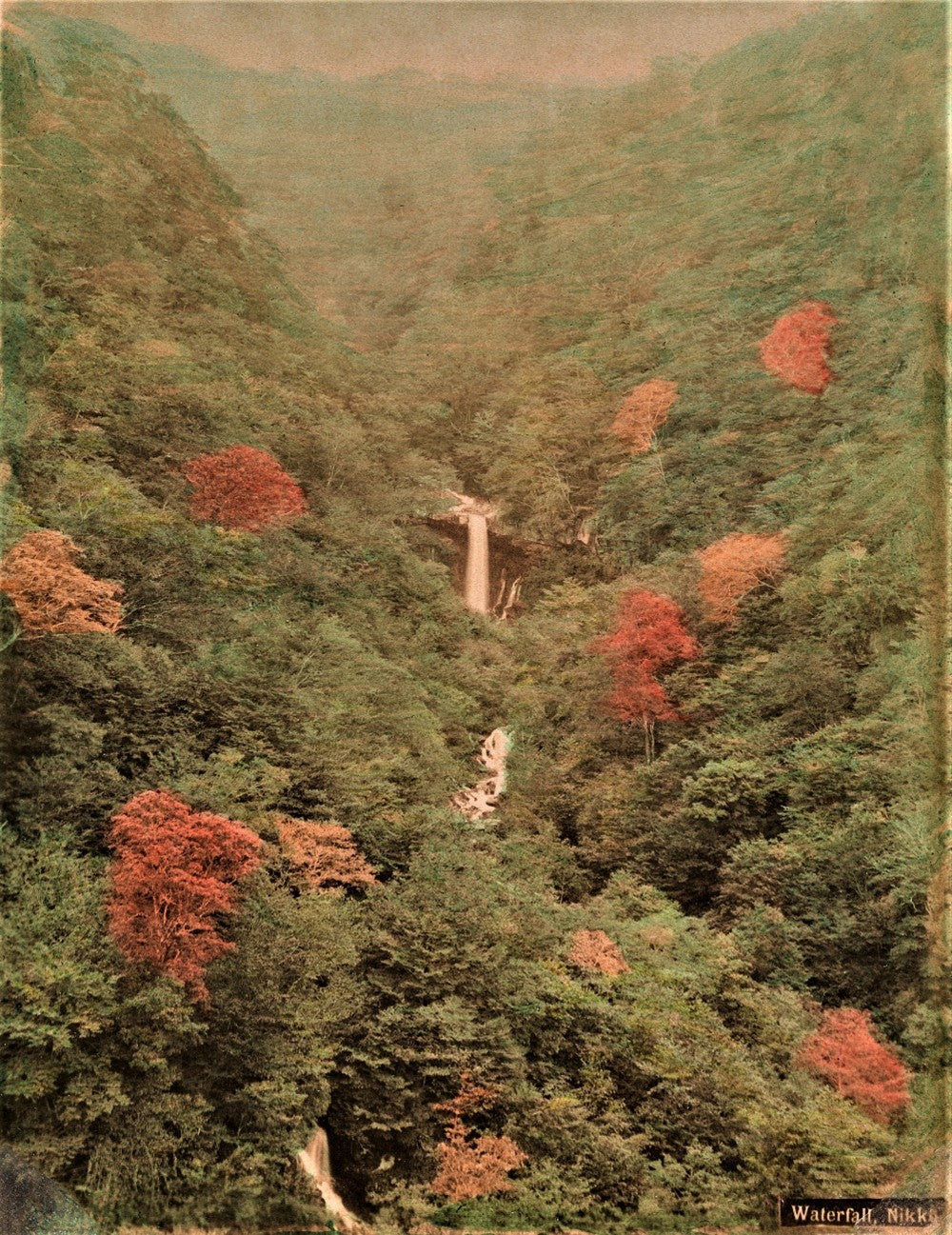 Hand-colored sepia photo of a waterfall in Nikko, Japan, surrounded by forested valley with autumn foliage