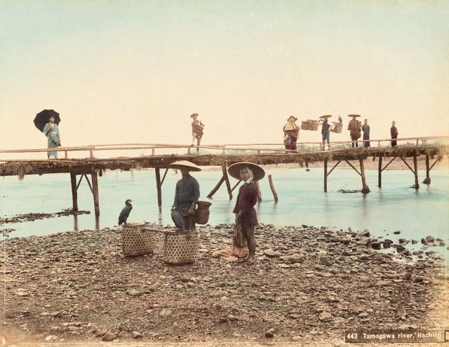 Hand-colored Meiji-era photograph of cormorant fishermen on the Tamagawa River with a wooden bridge and villagers in the background, Japan
