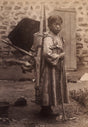 Sepia portrait of a young Korean figure wearing a traditional overcoat and carrying a bundle, photographed in a rustic courtyard during the early Korean Empire.
